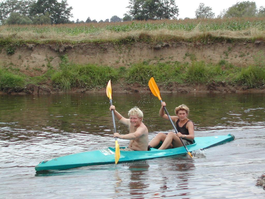 Two people paddle a green kayak on a calm river, using yellow paddles to move through the water.