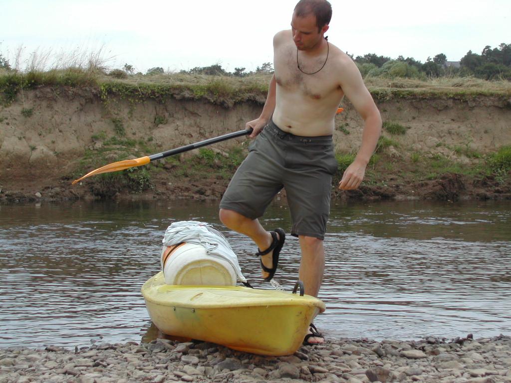 A man holding a paddle steps onto a yellow kayak loaded with gear on a rocky riverbank.