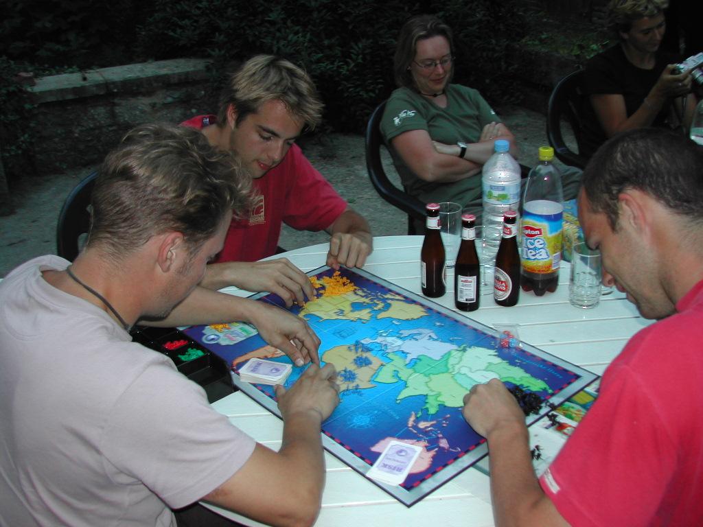 Three people play the board game Risk at an outdoor table, focusing on the game pieces and strategy.