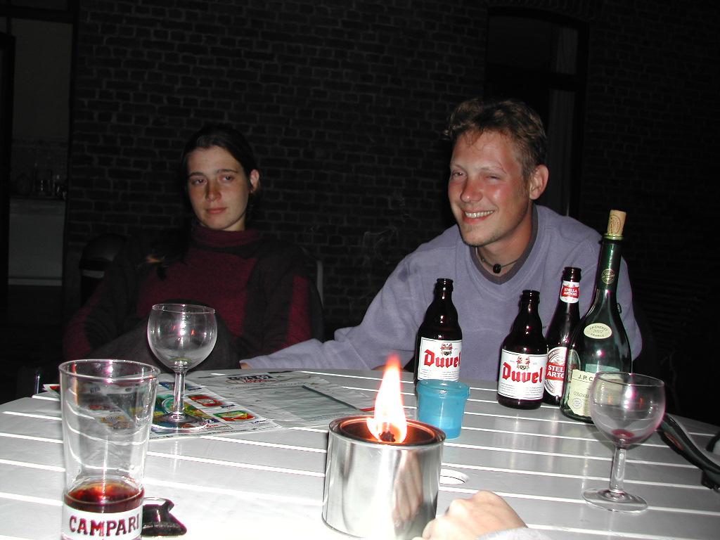 Two people sit at a table with drinks, a small open flame, and bottles of alcohol in front of them.