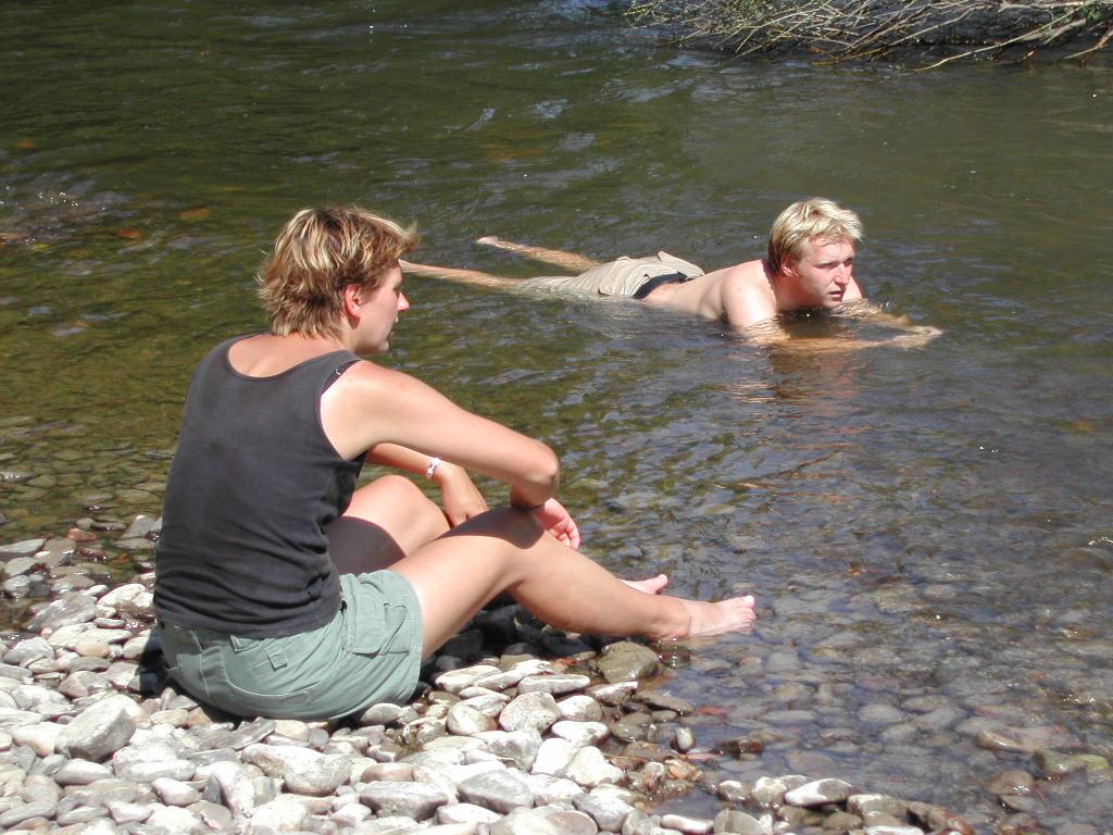 A woman sits on a rocky riverbank while a man lies in the shallow water, partially submerged.
