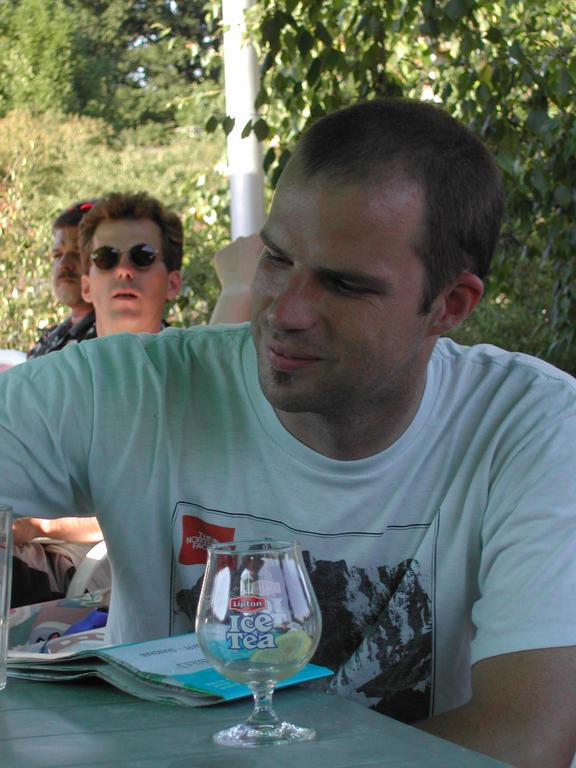A man in a white t-shirt sits at an outdoor table, smiling, with a glass of iced tea nearby.