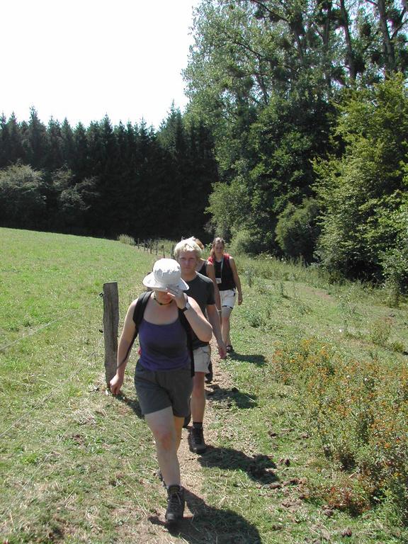 Three people walk along a narrow dirt path in a grassy area, wearing backpacks and casual outdoor clothing.