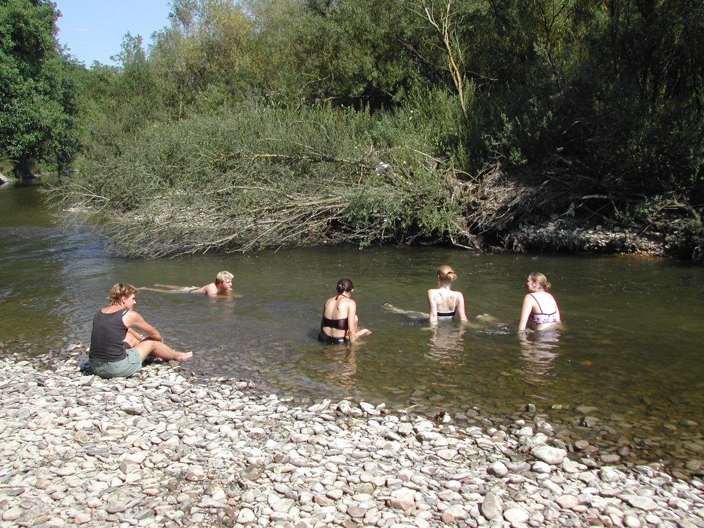 Four people sit and swim in a shallow river, while another person sits on the rocky shore watching.