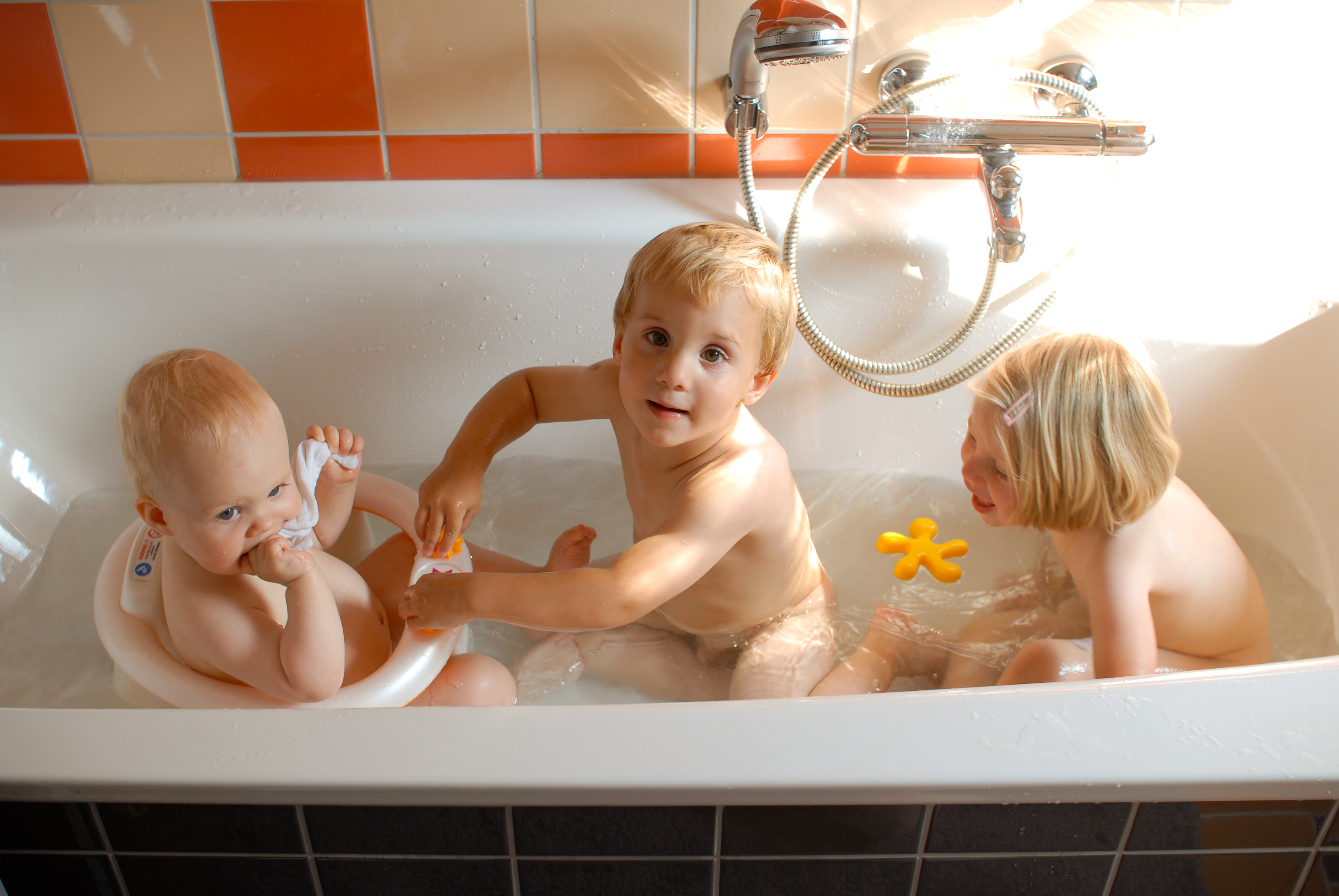 Three young children are playing together in a bathtub, splashing water and holding bath toys.