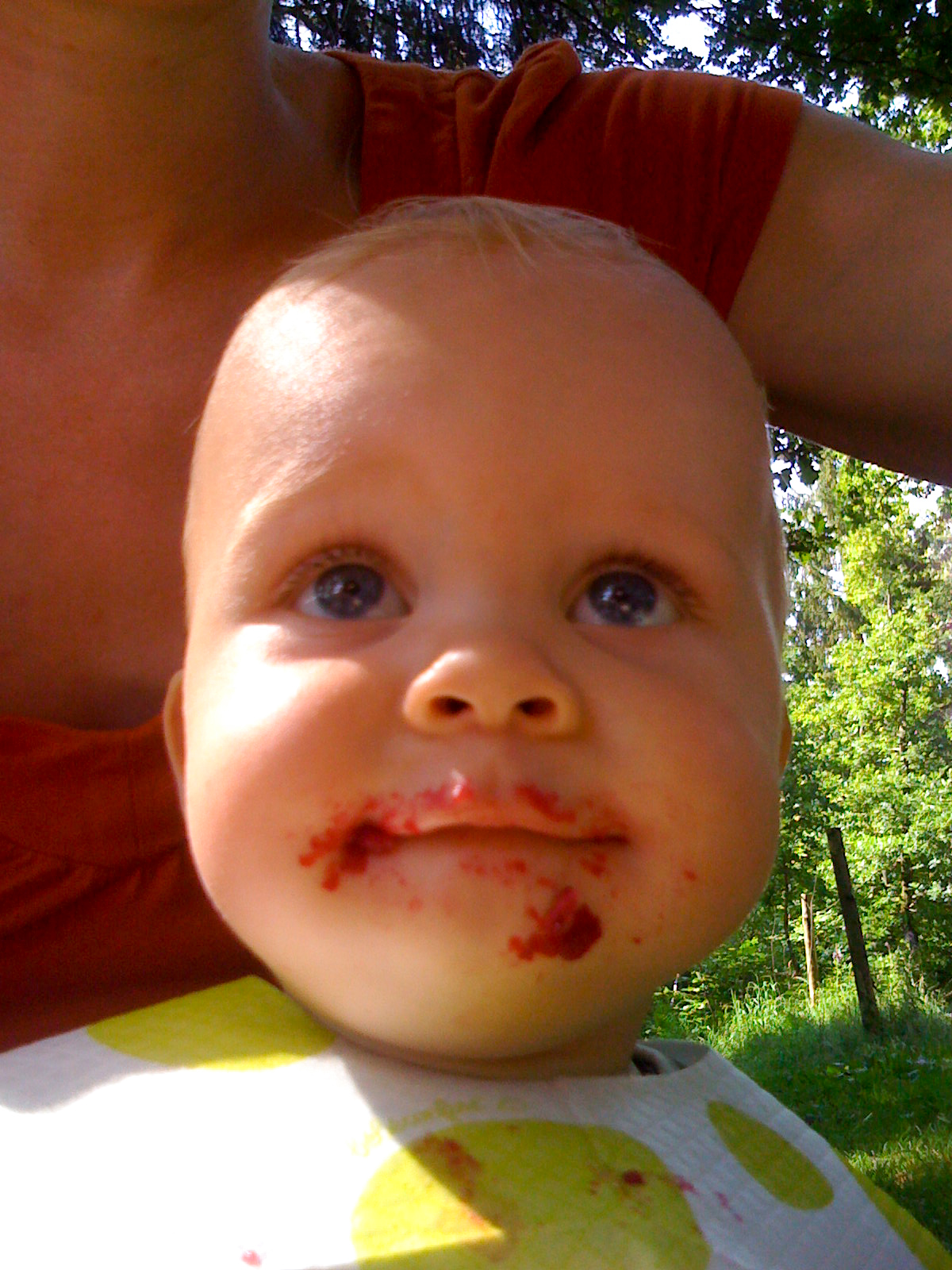 A baby with food smeared on their face sits outside, wearing a bib, with an adult partially visible behind them.
