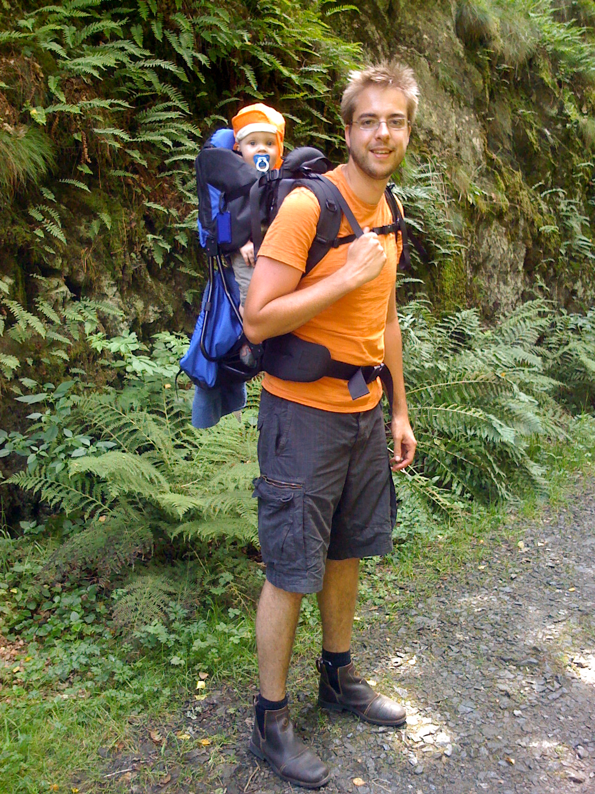 A man wearing an orange shirt hikes on a forest trail, carrying a baby in a backpack carrier.