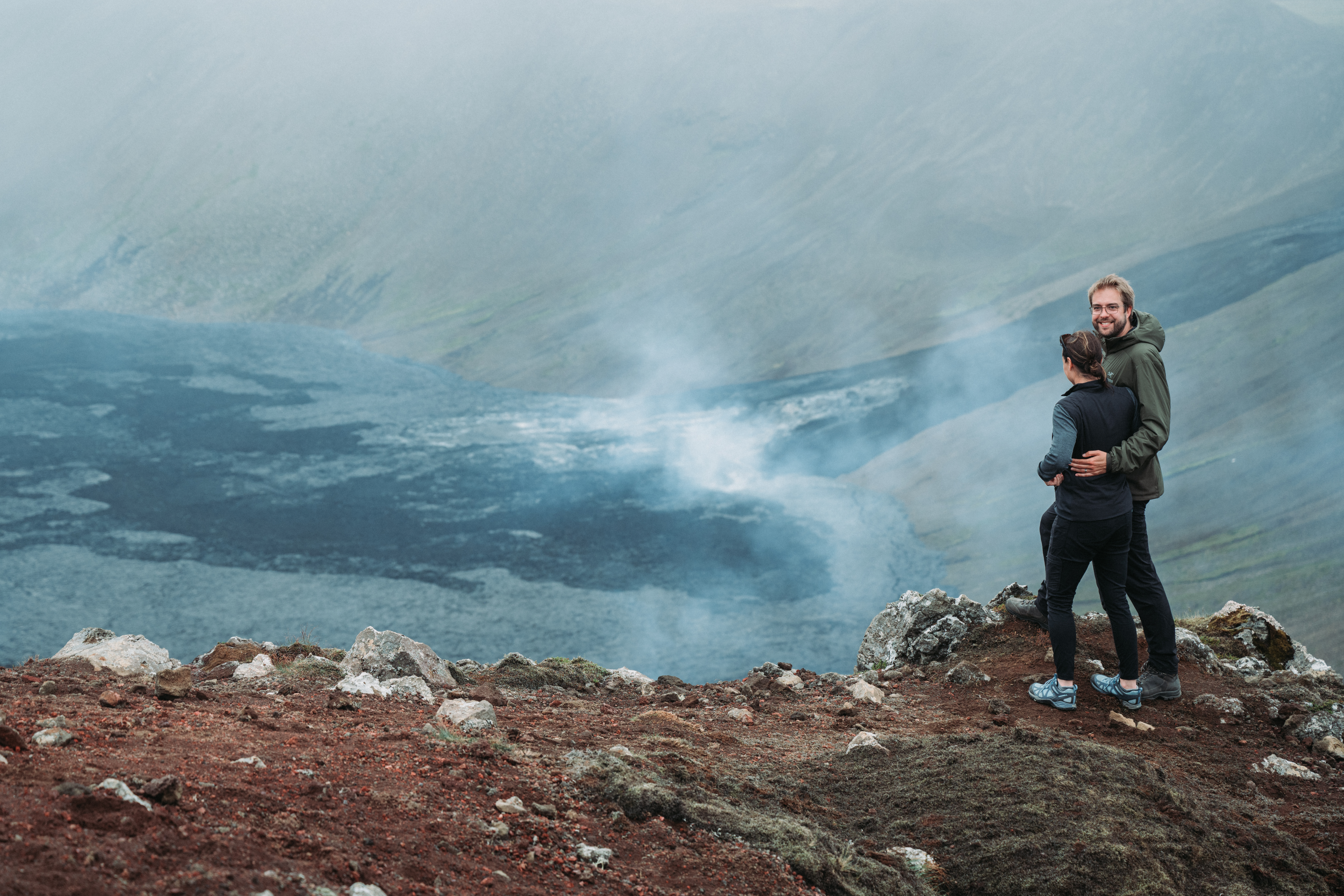 Two people stand on a rocky hill, overlooking a steaming volcanic landscape in Gerlingadalur, Iceland.