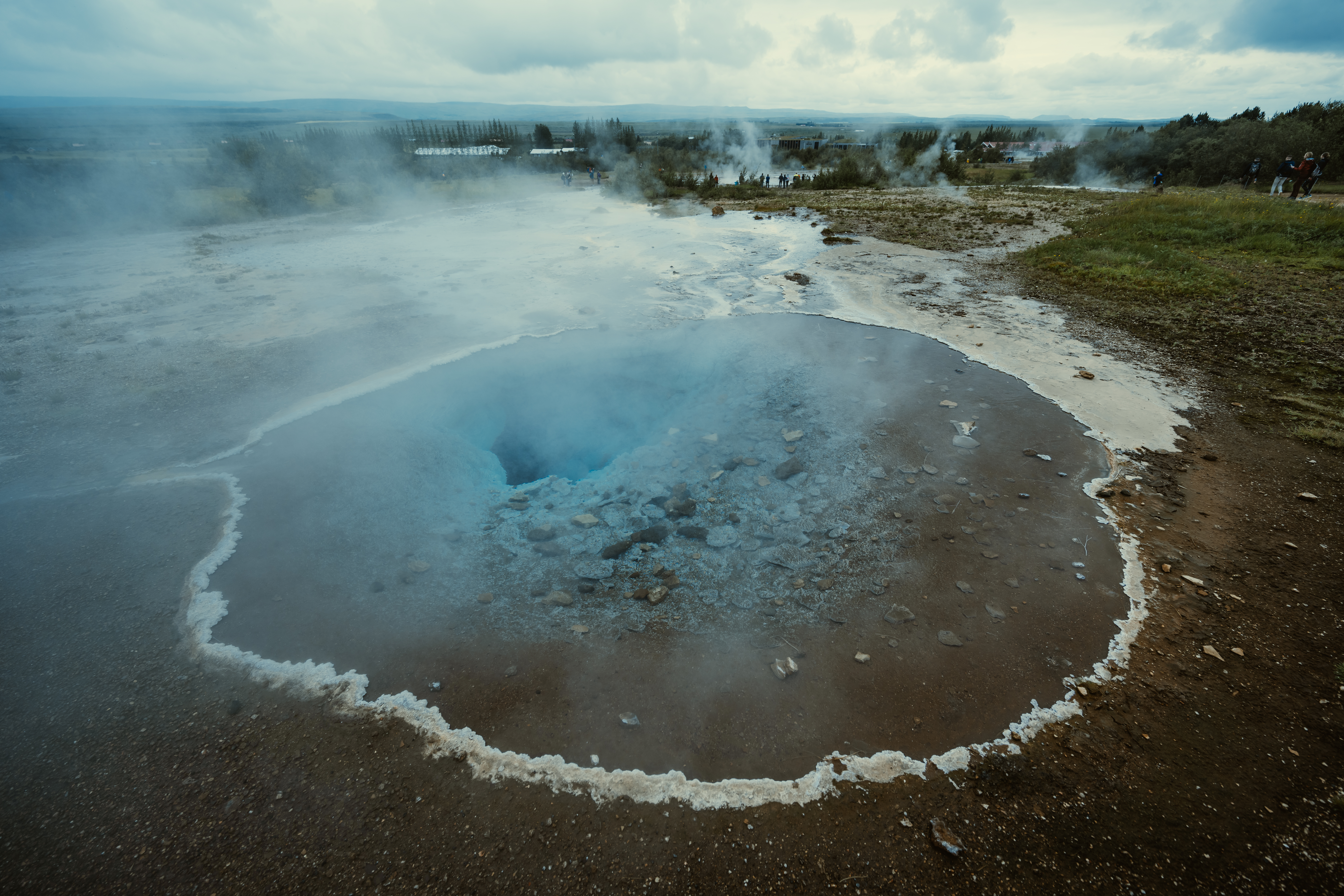 A steaming hot spring with a deep blue center at Geysir, Iceland, surrounded by mineral deposits and rocky ground.