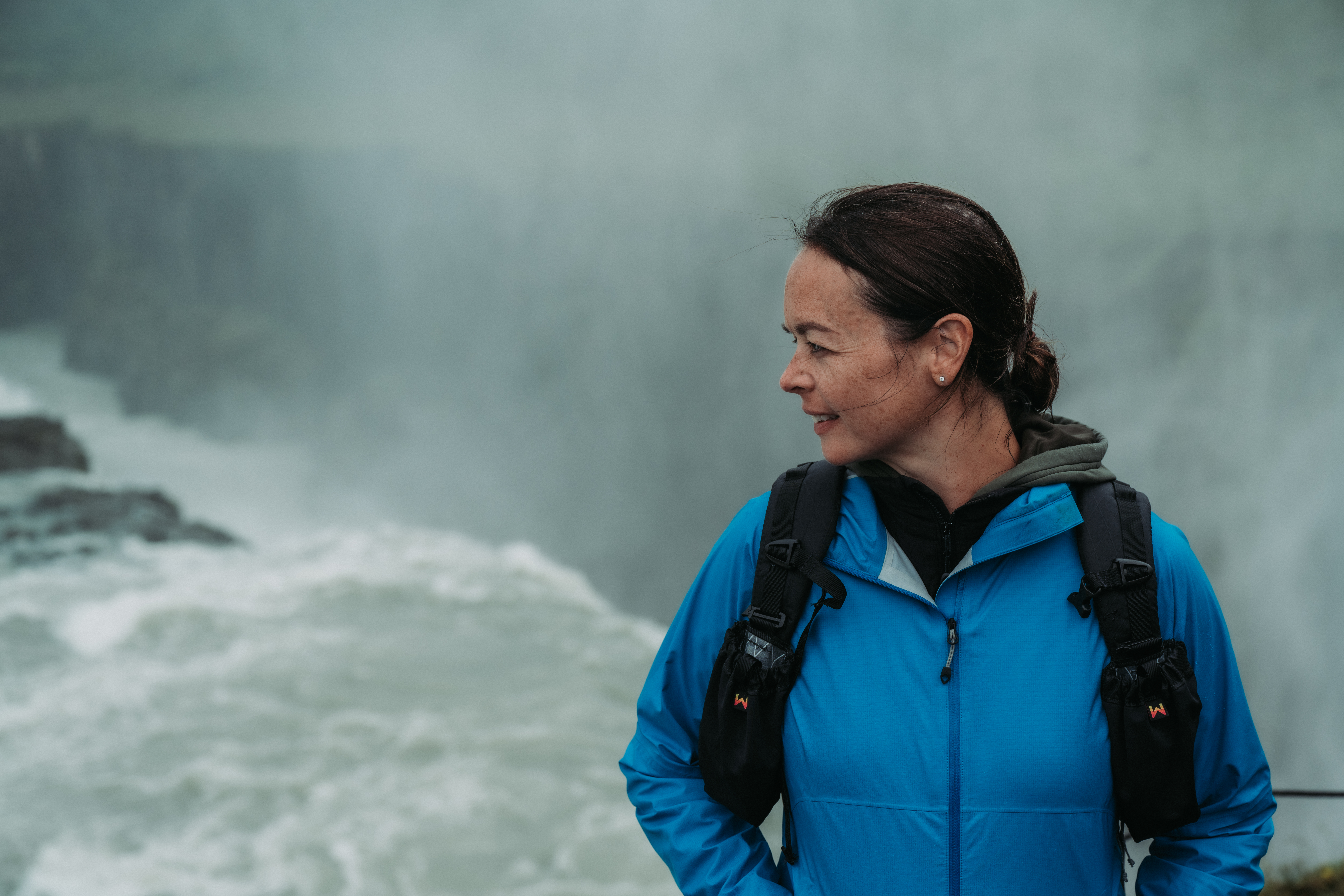 A person wearing a blue jacket and backpack stands near Gullfoss Falls, looking to the side.