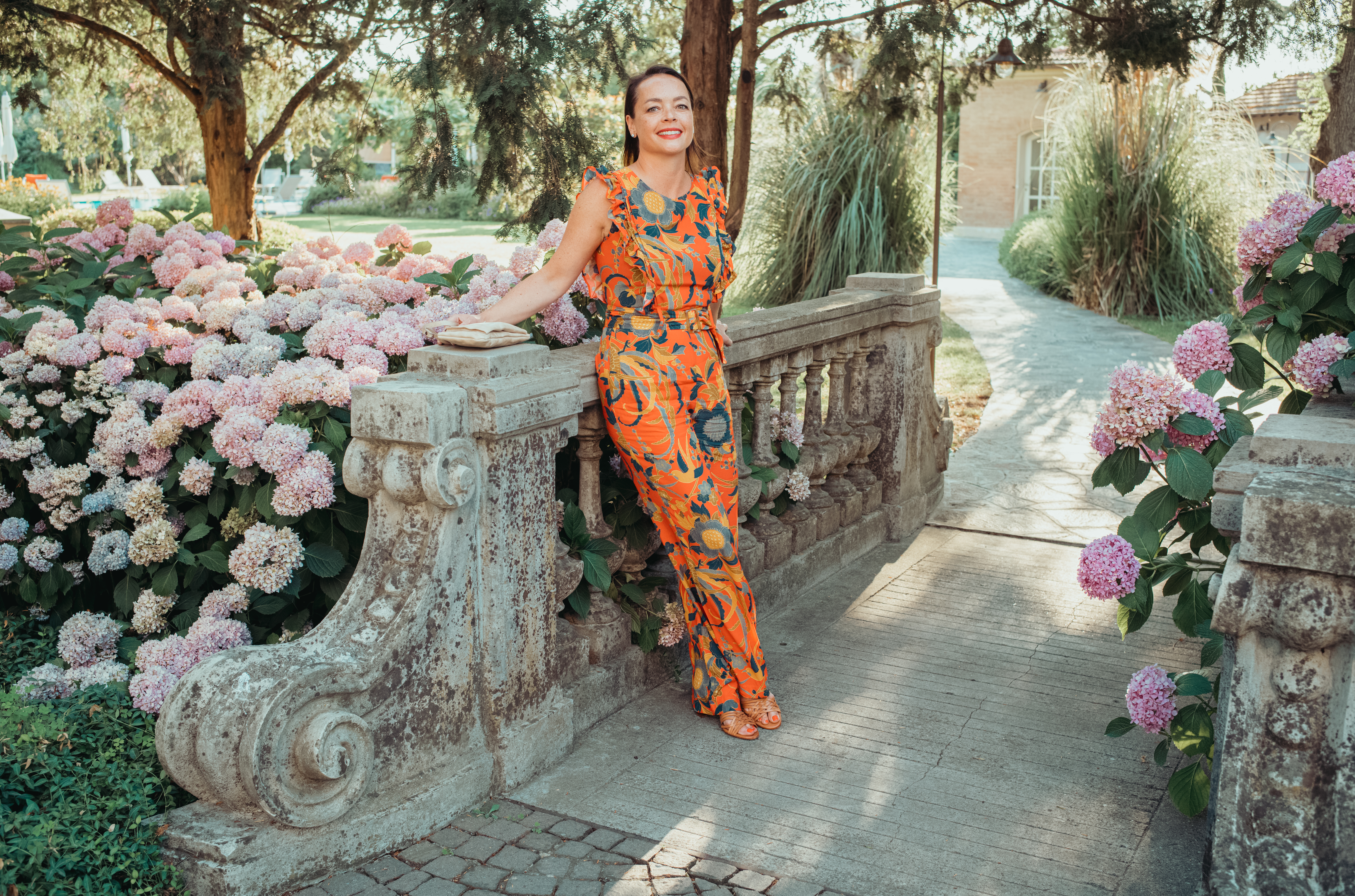 A woman in a colorful outfit leans on a stone railing, smiling, surrounded by pink flowers and greenery.