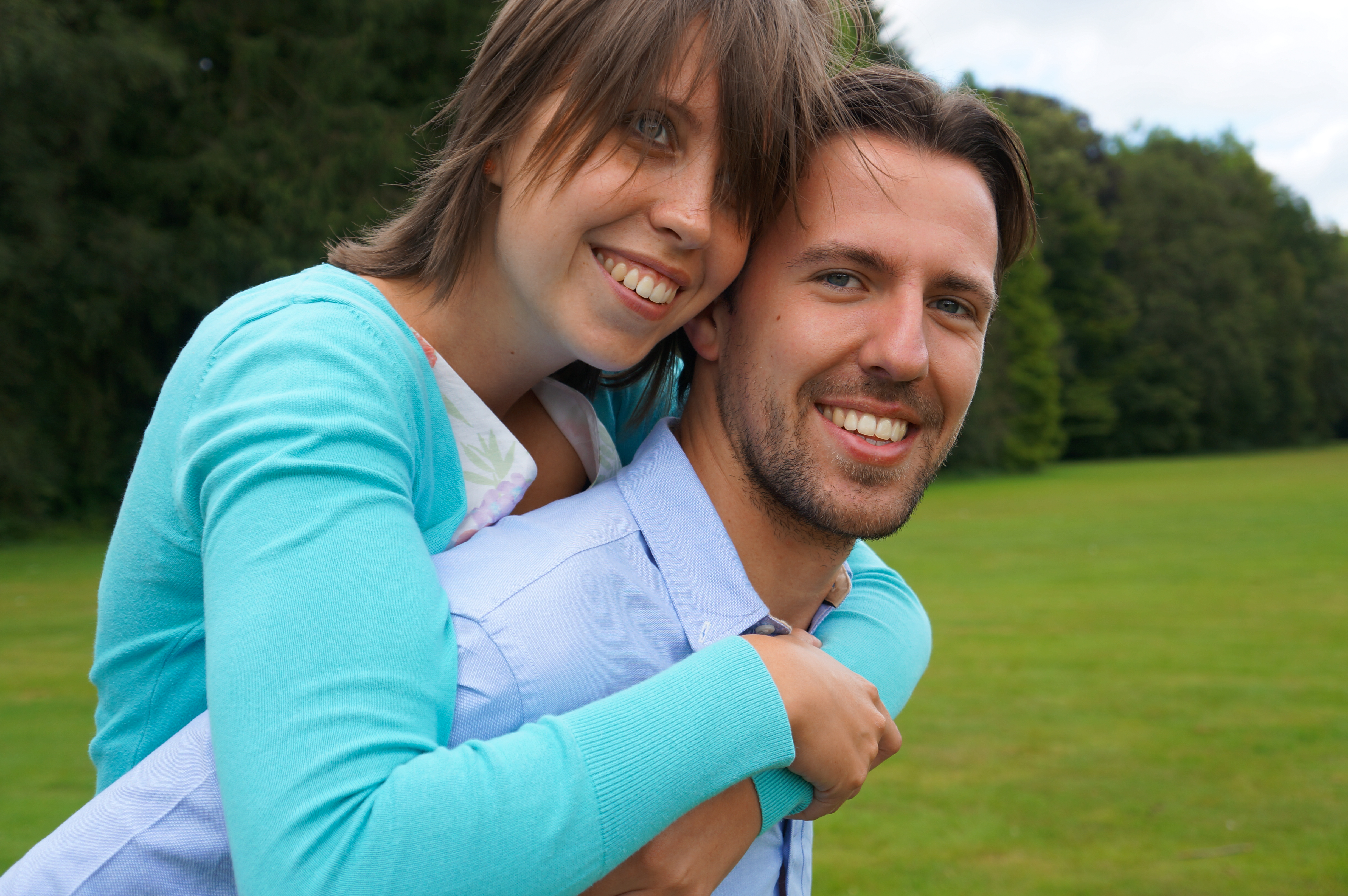 A smiling woman in a turquoise sweater hugs a smiling man from behind in an outdoor setting.