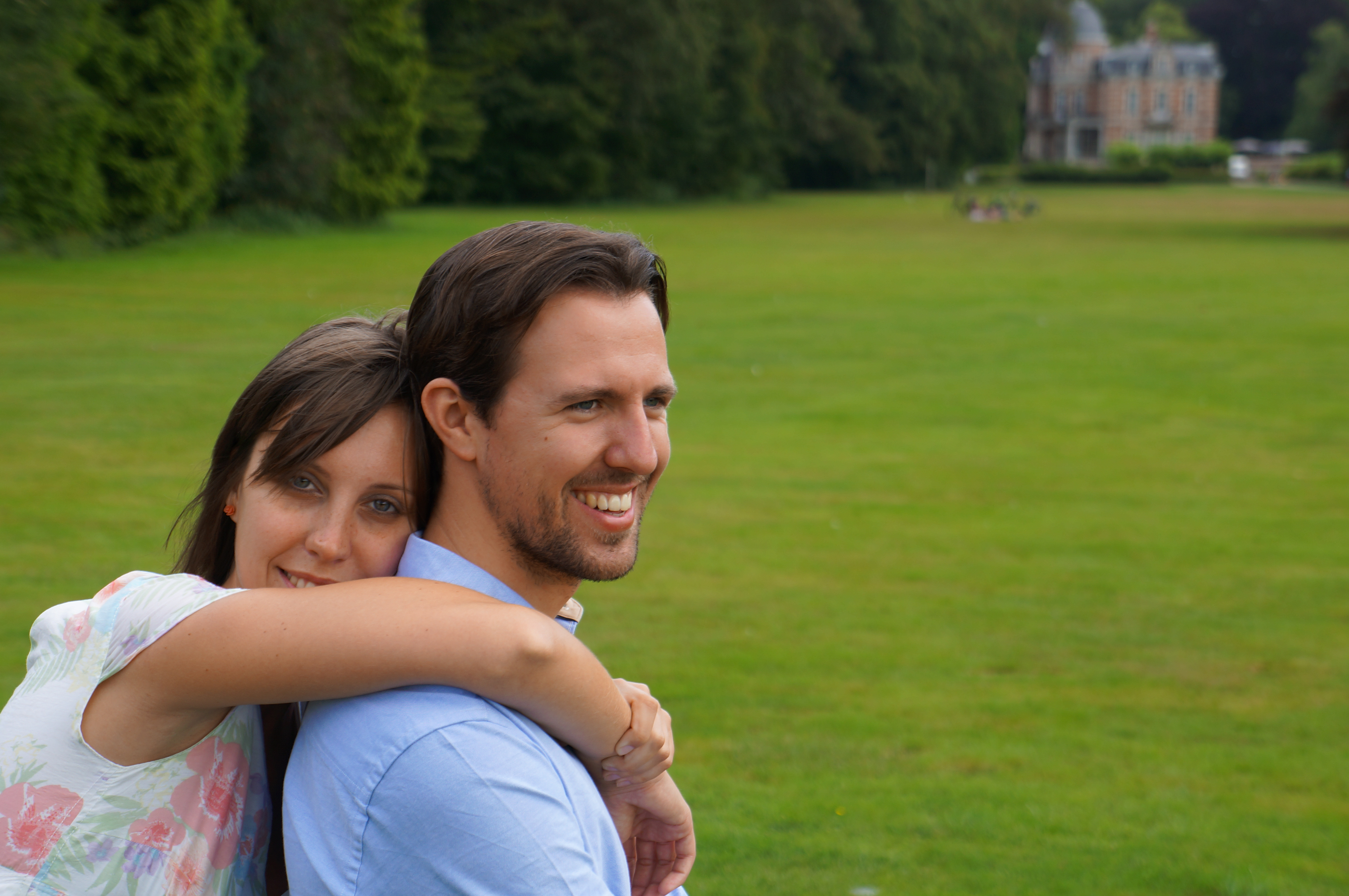 A woman hugs a smiling man from behind while standing in a grassy field with trees and a building in the background.