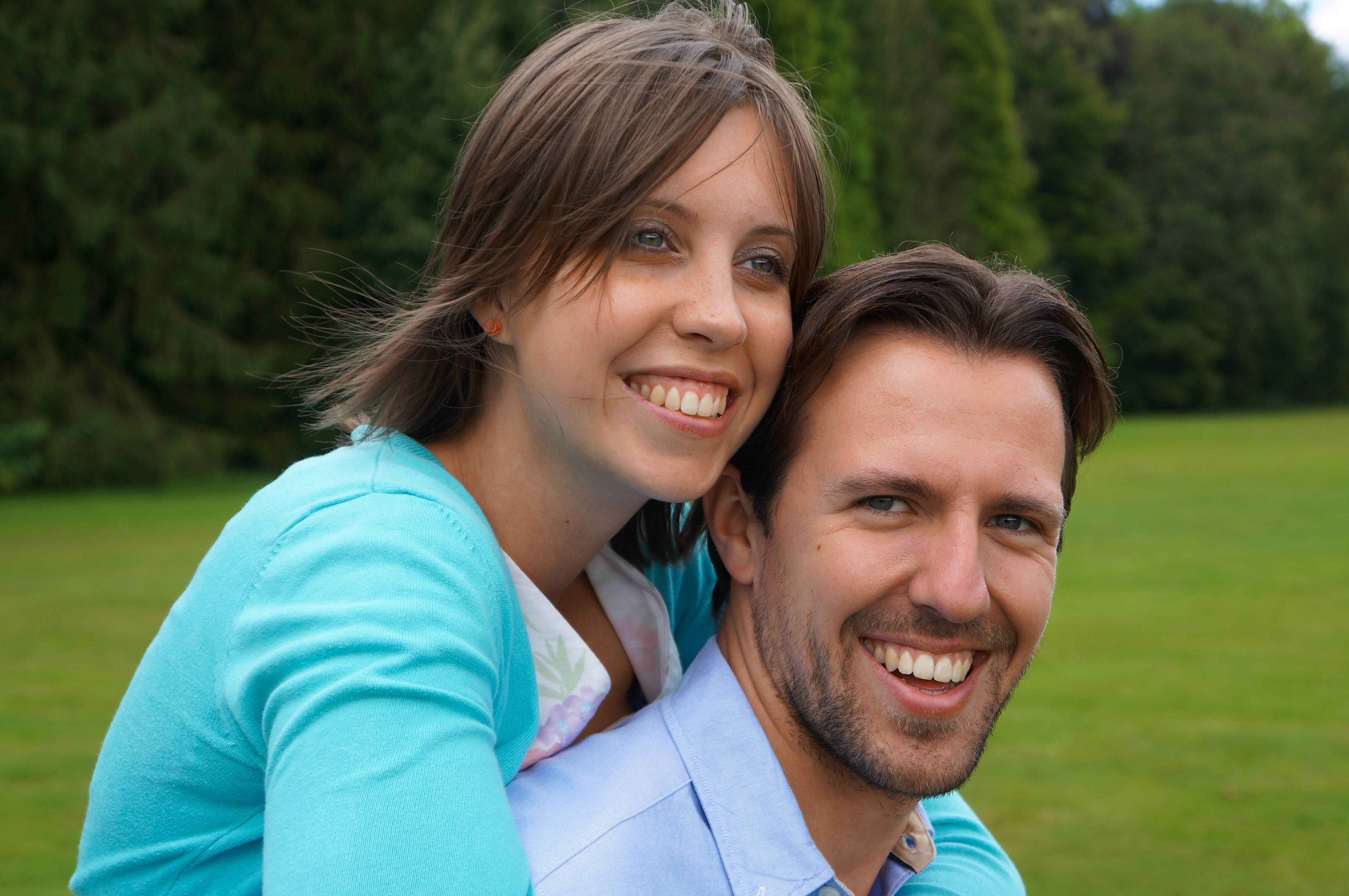 A smiling woman in a light blue sweater hugs a smiling man from behind in an outdoor setting.