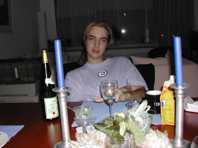 A person sits at a dinner table holding a wine glass, with candles, a bottle, and food items nearby.