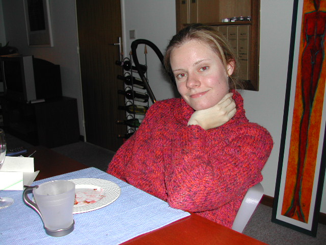 A woman in a red sweater sits at a table with an empty plate, smiling at the camera.