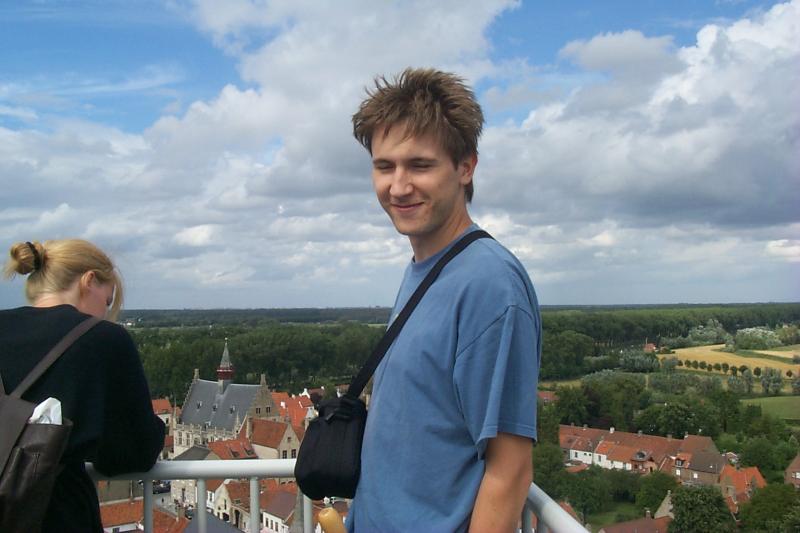 A man in a blue shirt smiles at the camera while standing on a high viewpoint with a scenic background.
