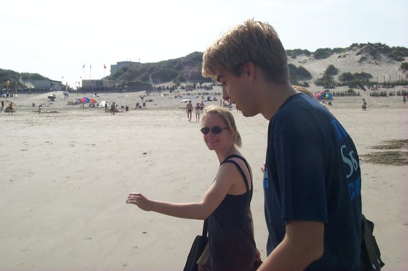 Two people walk along a sandy beach, one smiling and wearing sunglasses.
