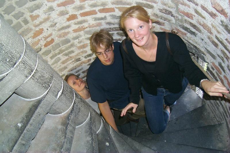 Three people climbing a narrow spiral staircase with brick walls, one smiling at the camera while reaching for the railing.