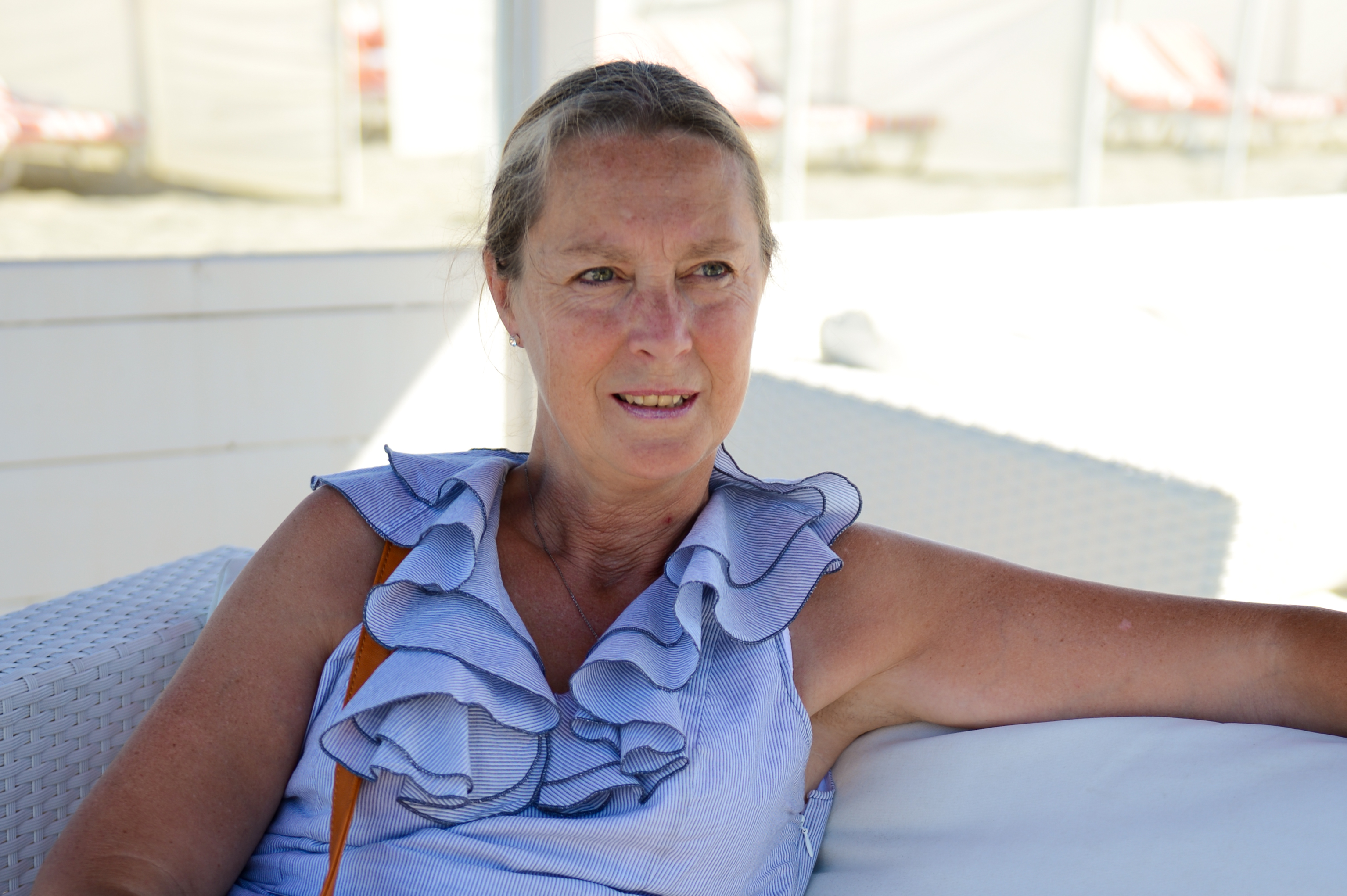 A woman sits on a white outdoor couch, wearing a blue ruffled top and looking slightly to the side.