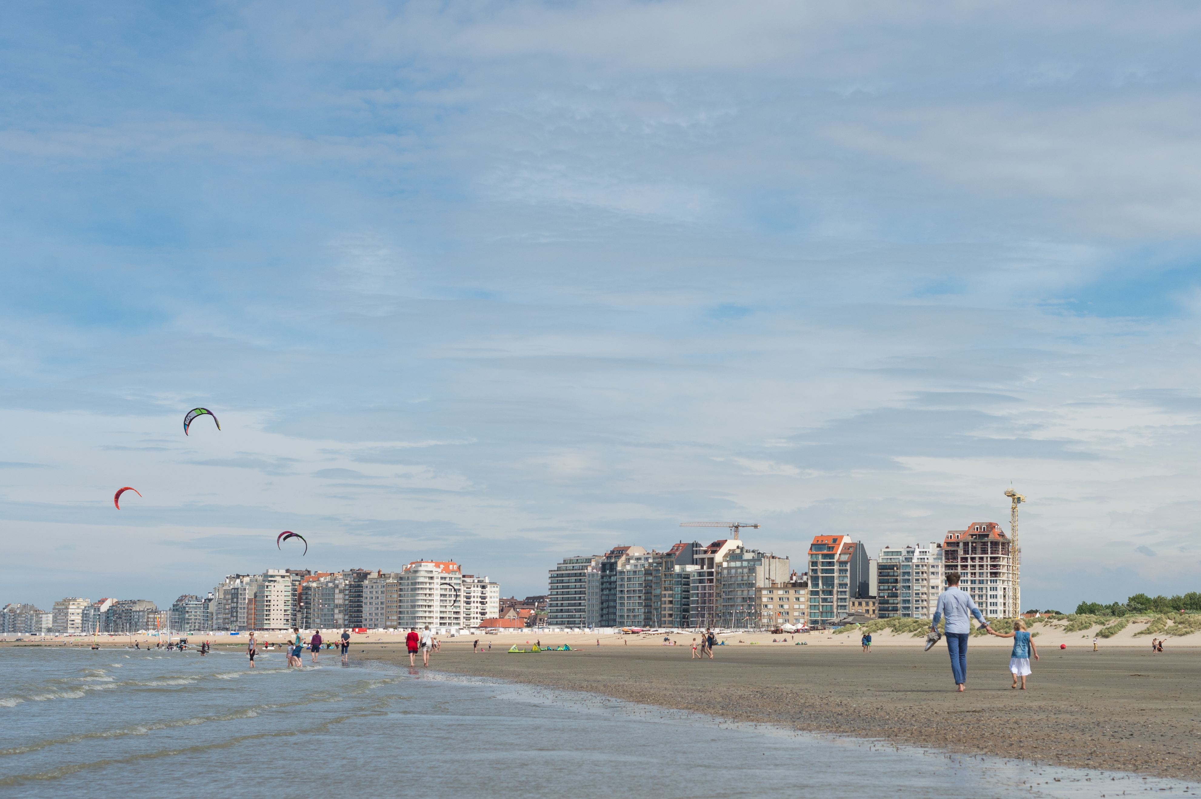 A man and a child walk barefoot on the beach, while people and kite surfers enjoy the shoreline.
