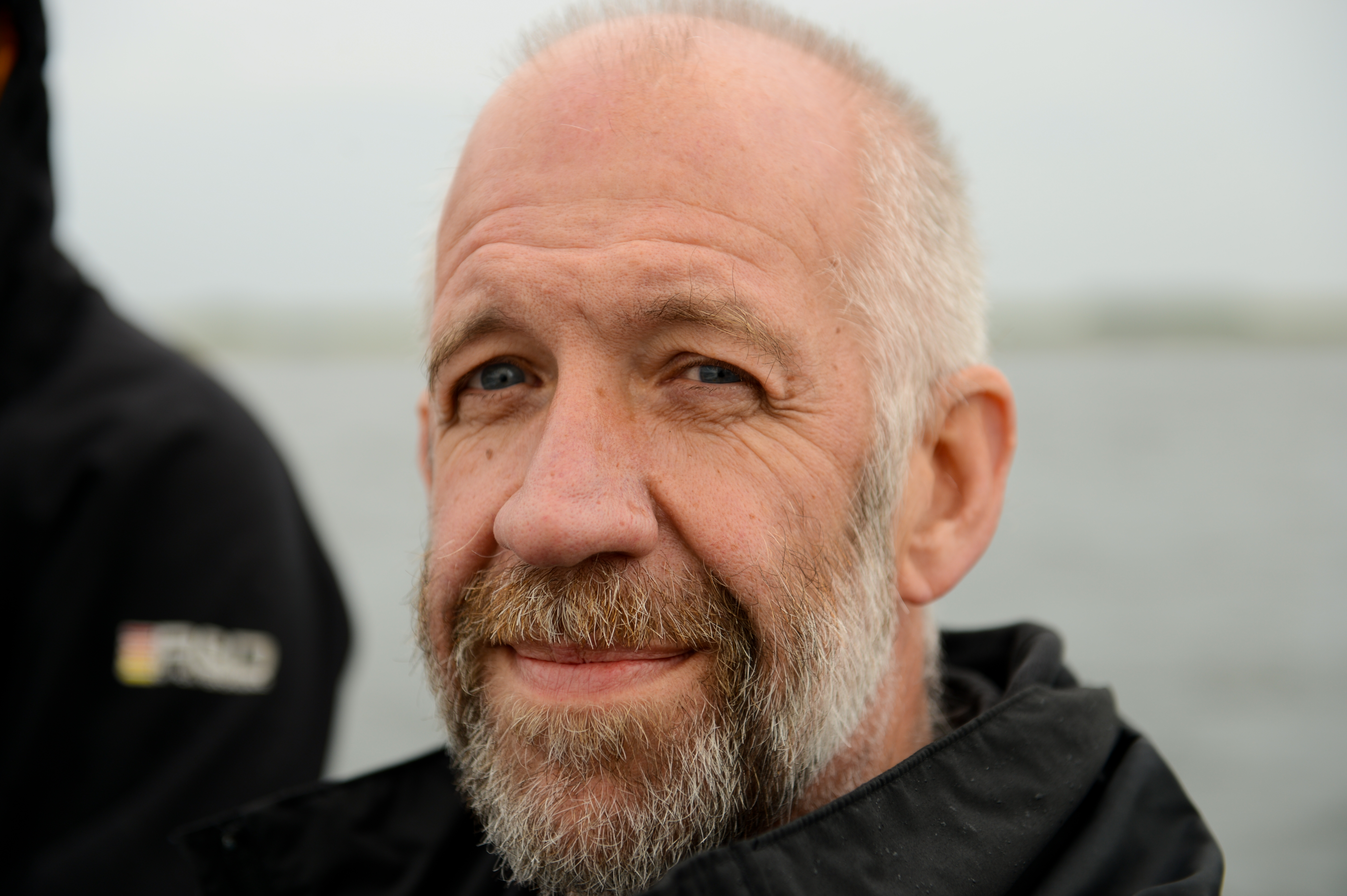 A bearded man in a black jacket smiles slightly while looking at the camera, with water in the background.