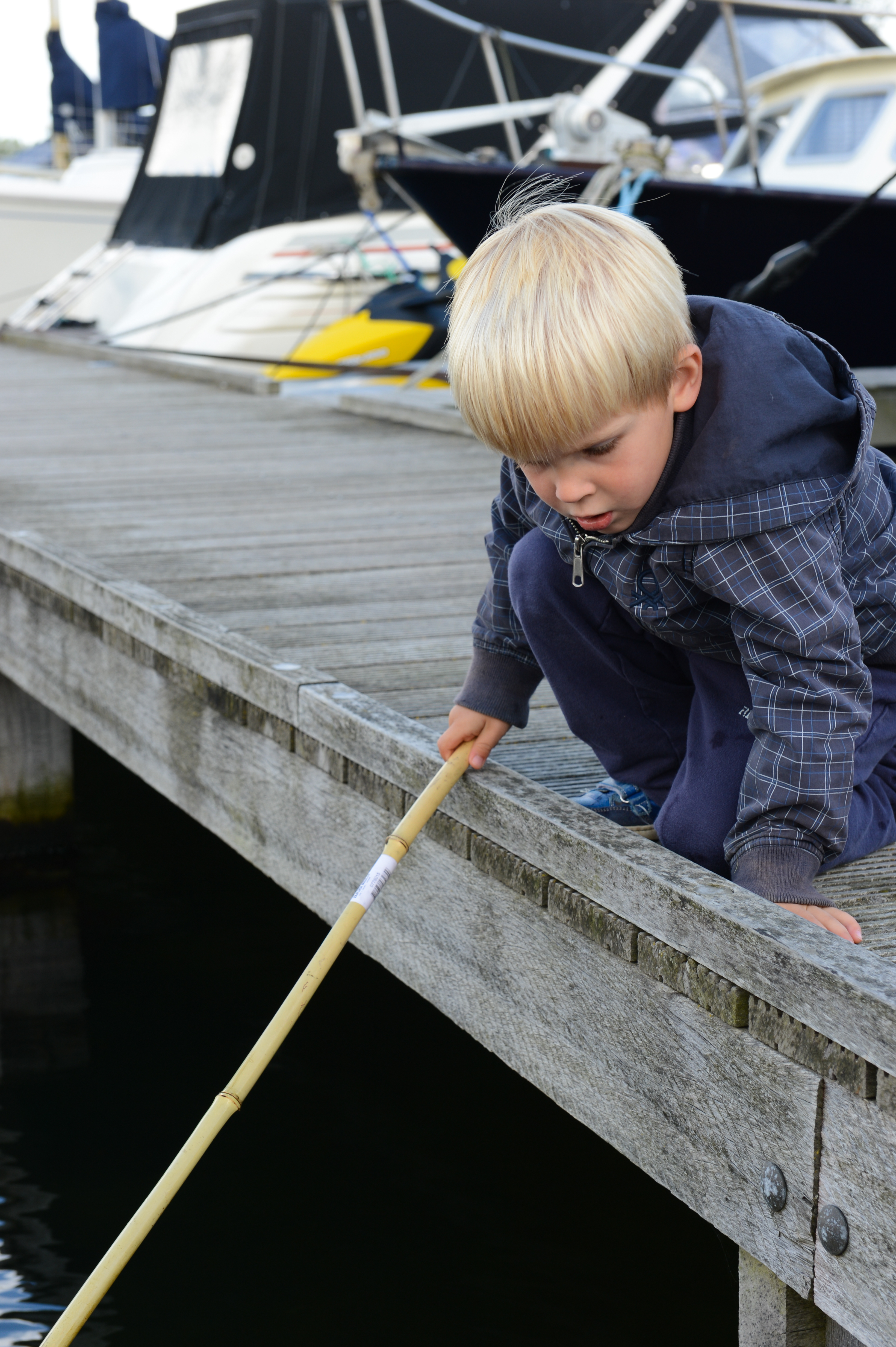 A young child crouches on a wooden dock, holding a long stick over the water, looking down intently.
