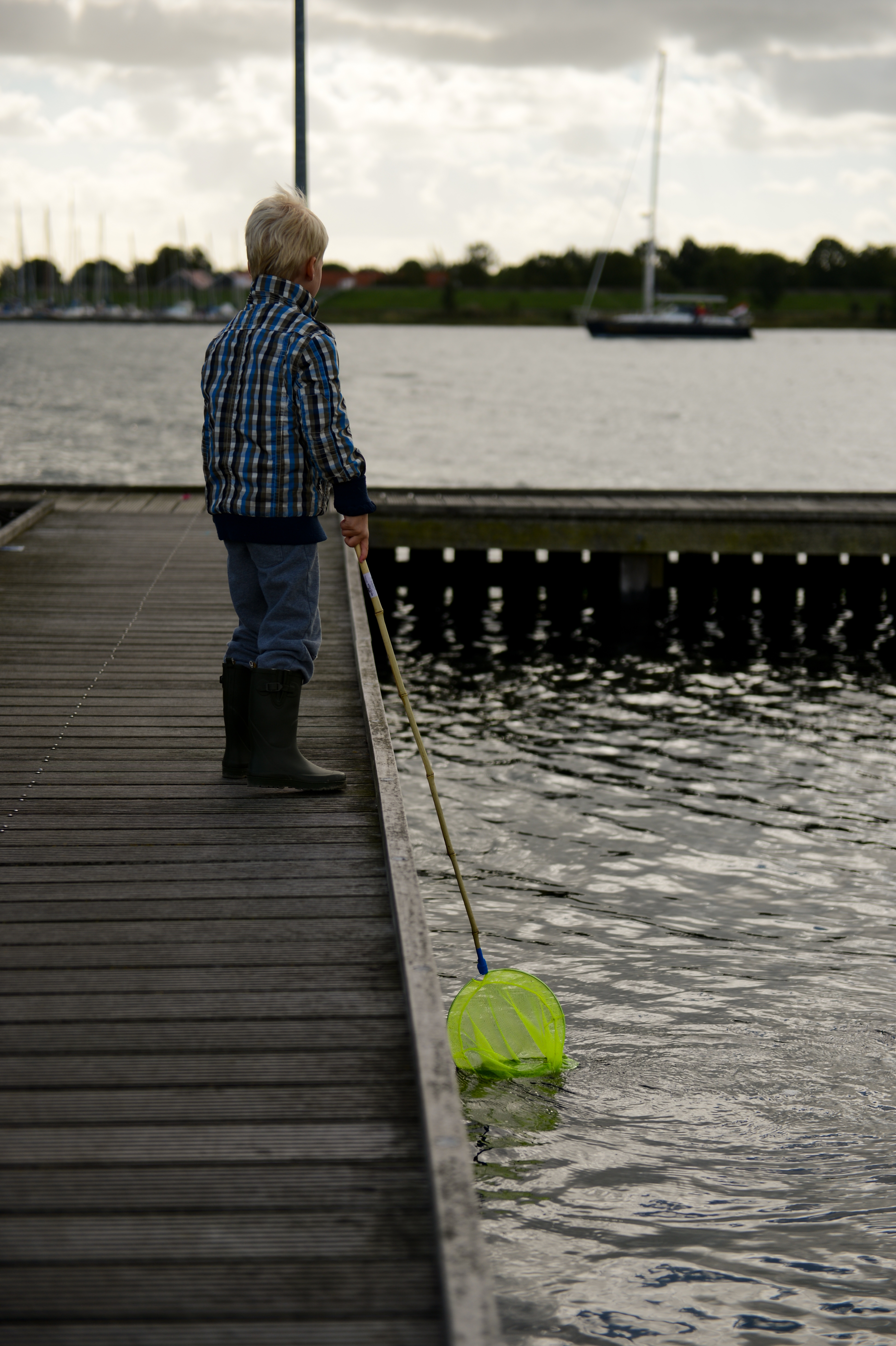 A child stands on a wooden dock, dipping a green fishing net into the water.