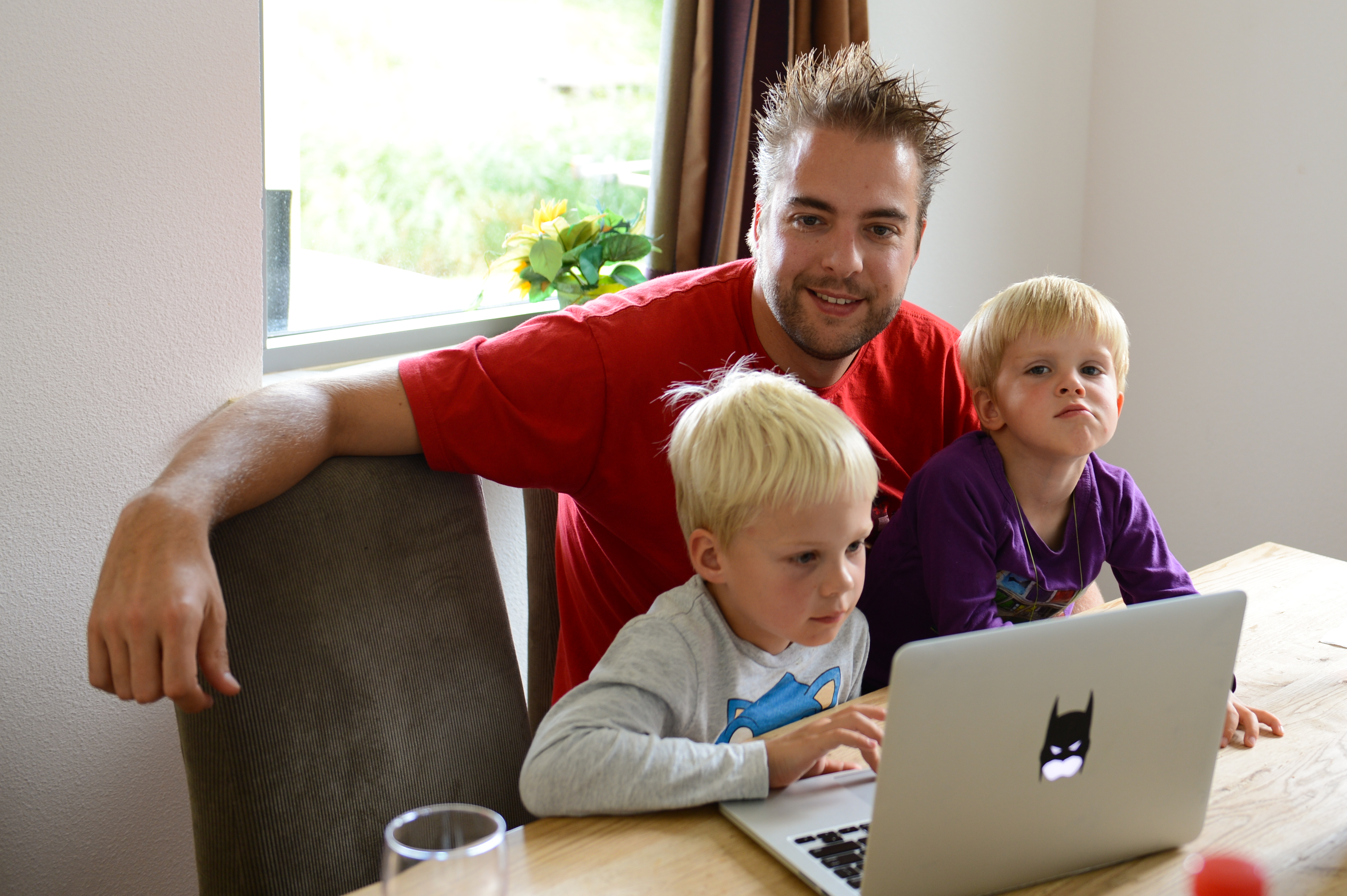 A man and two young children sit at a table, looking at a laptop screen together.