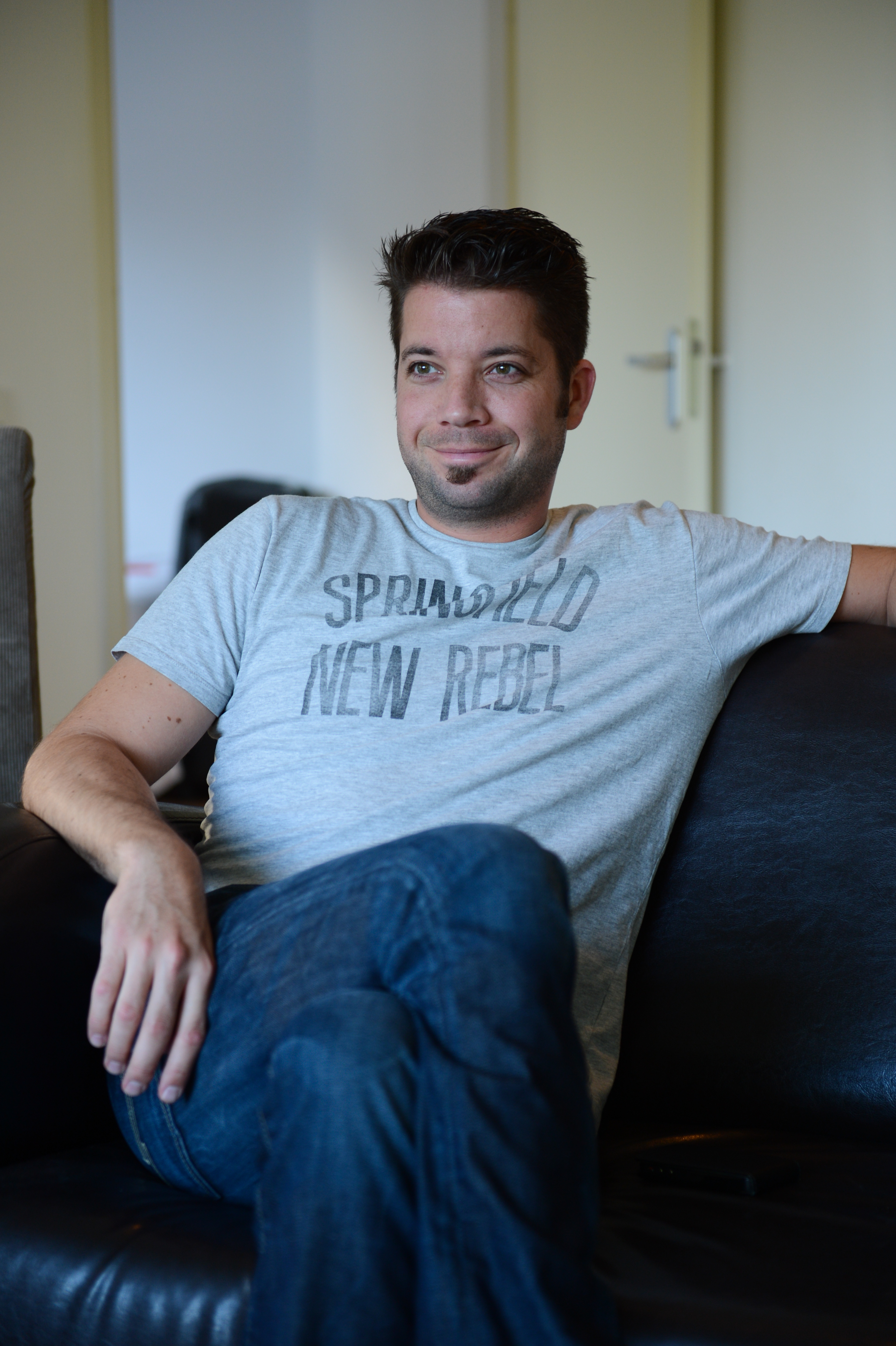 A man in a gray t-shirt sits on a black couch, smiling and looking off to the side.