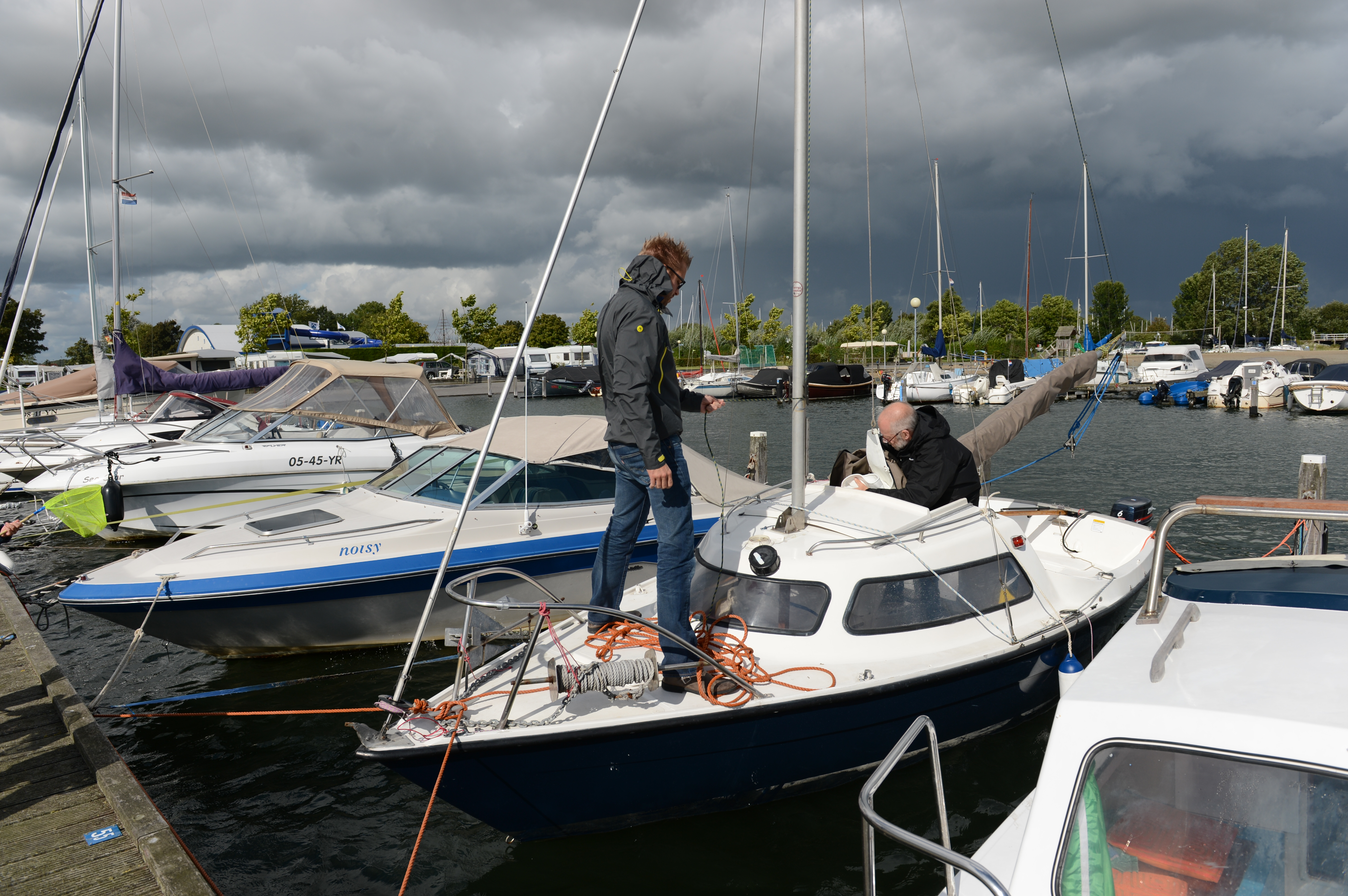Two people working on a small sailboat in a marina, one standing on the deck and the other seated.
