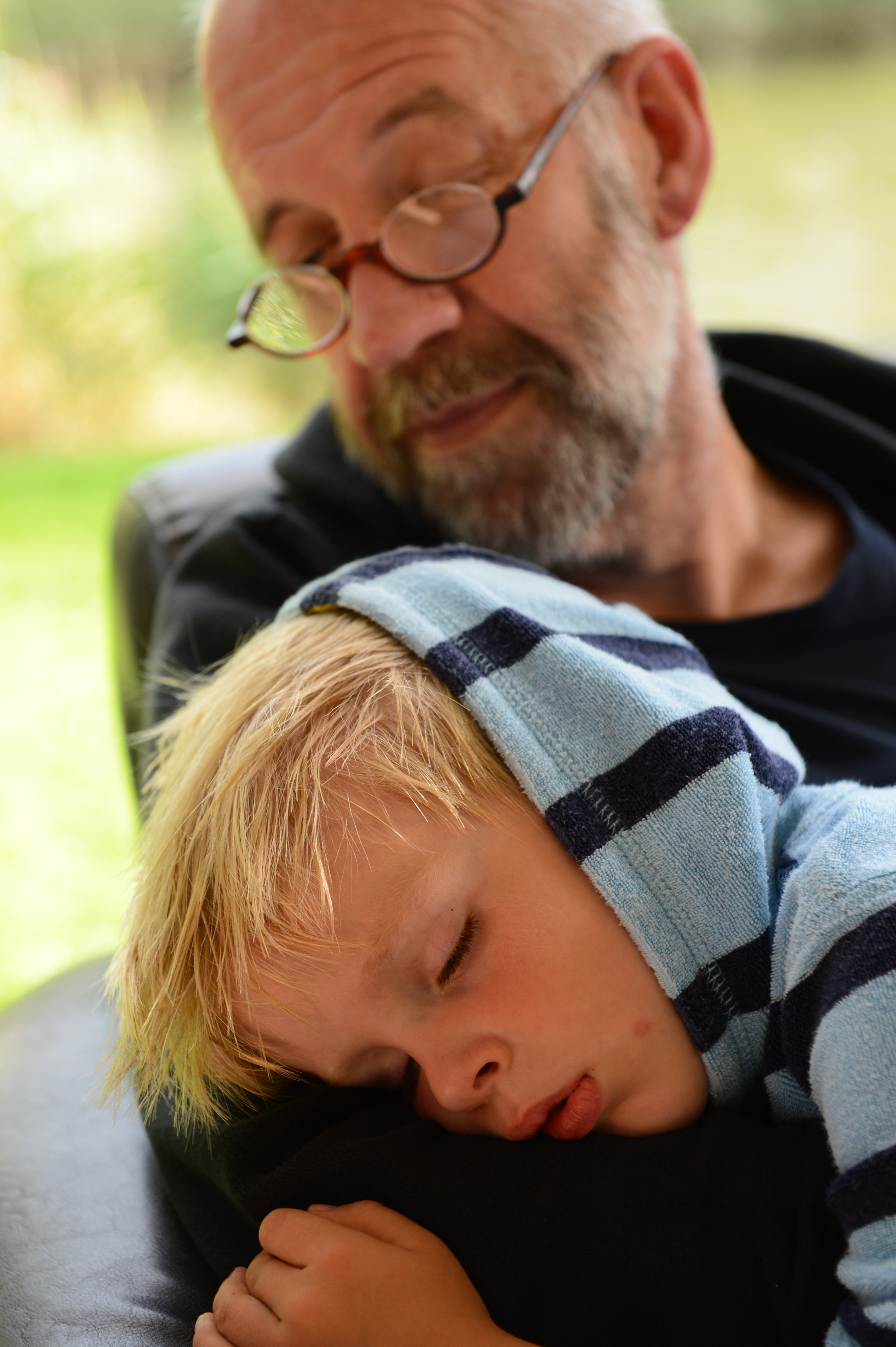 A child in a striped hoodie sleeps on an older man's arm while he looks down with a gentle expression.