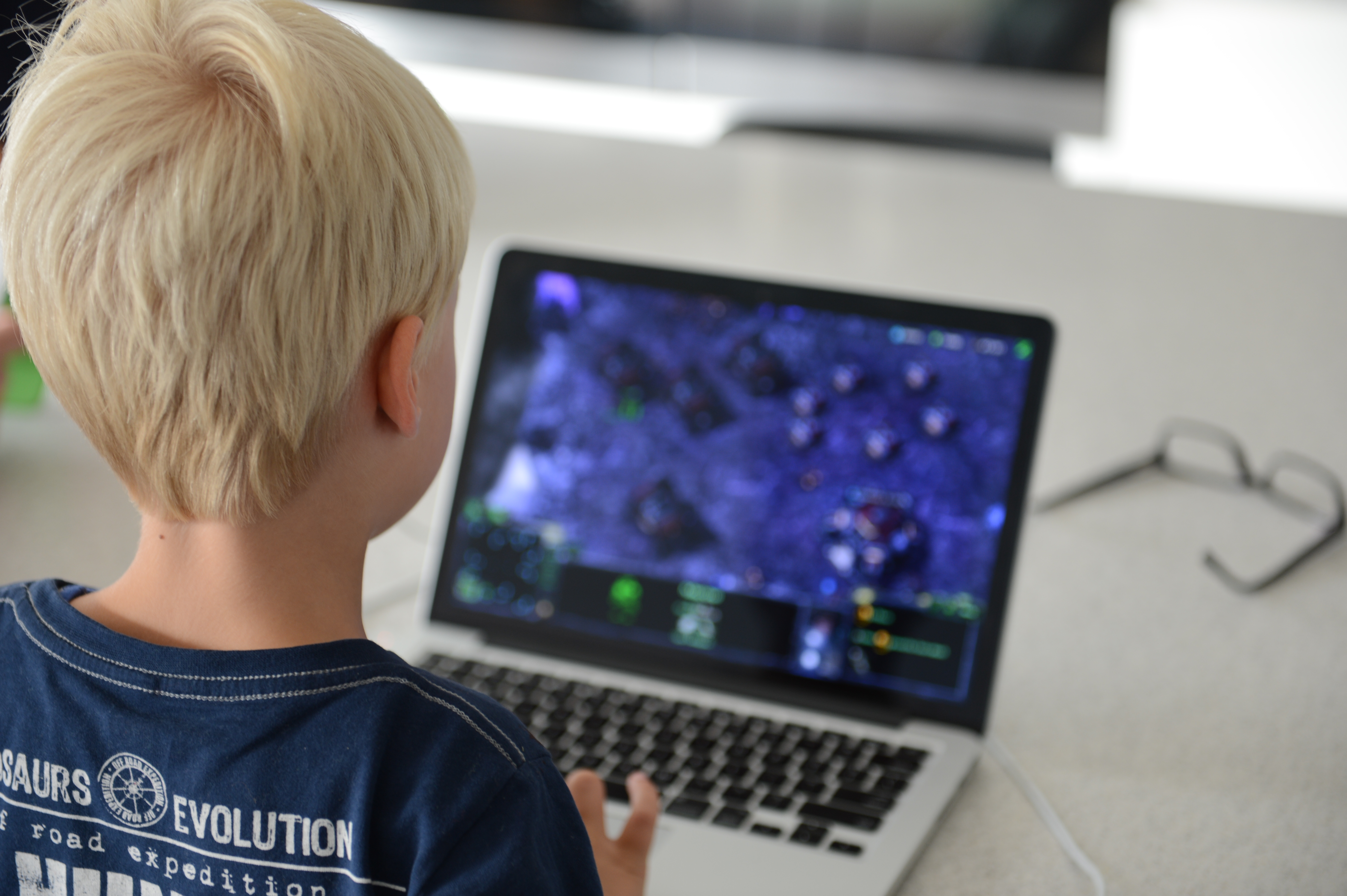 A child with blonde hair plays a strategy video game on a laptop at a table.