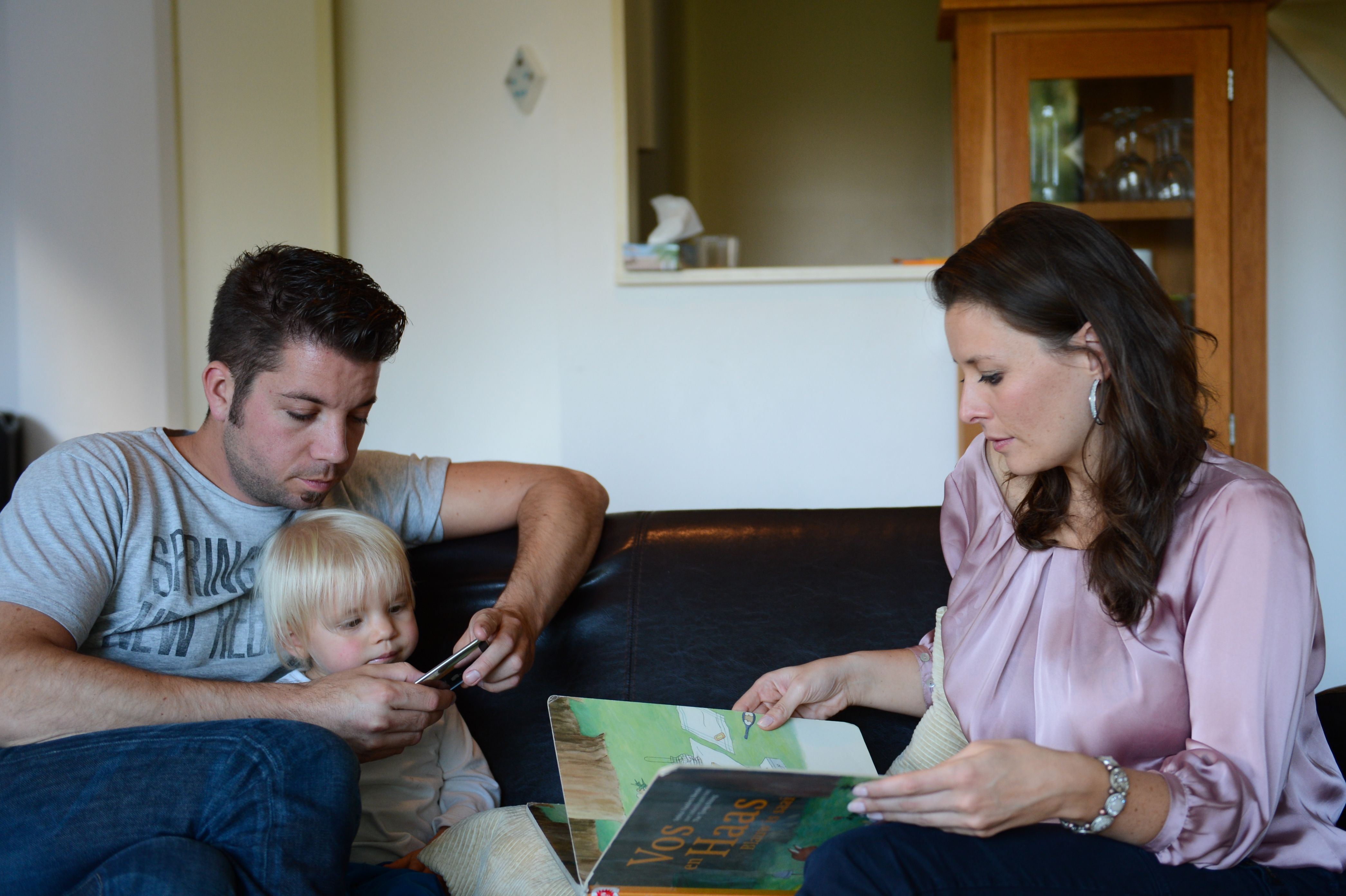 A woman reads a book on a couch while a man and child look at a phone together.