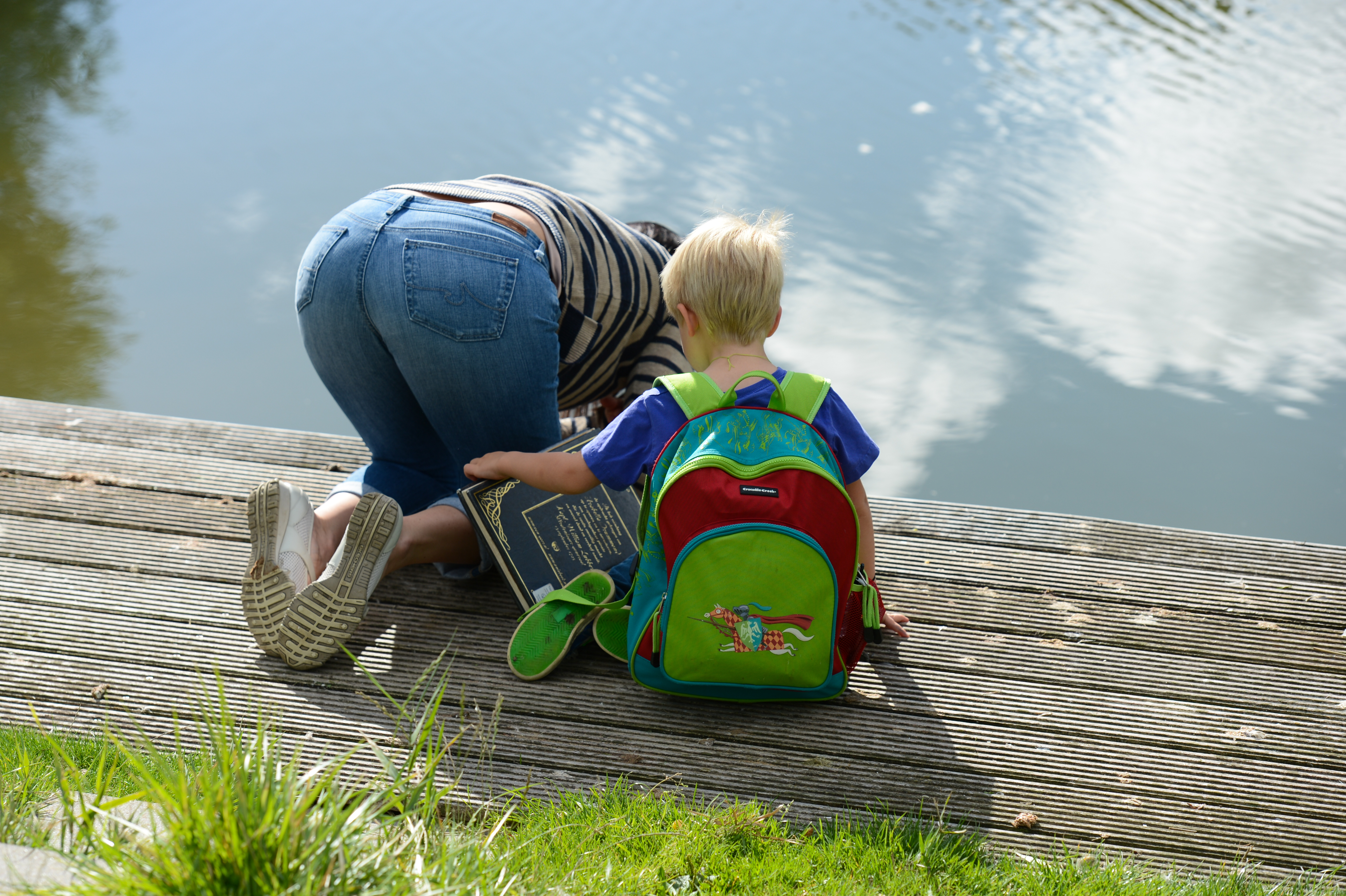 A child with a colorful backpack sits on a wooden dock, watching an adult leaning over the water.