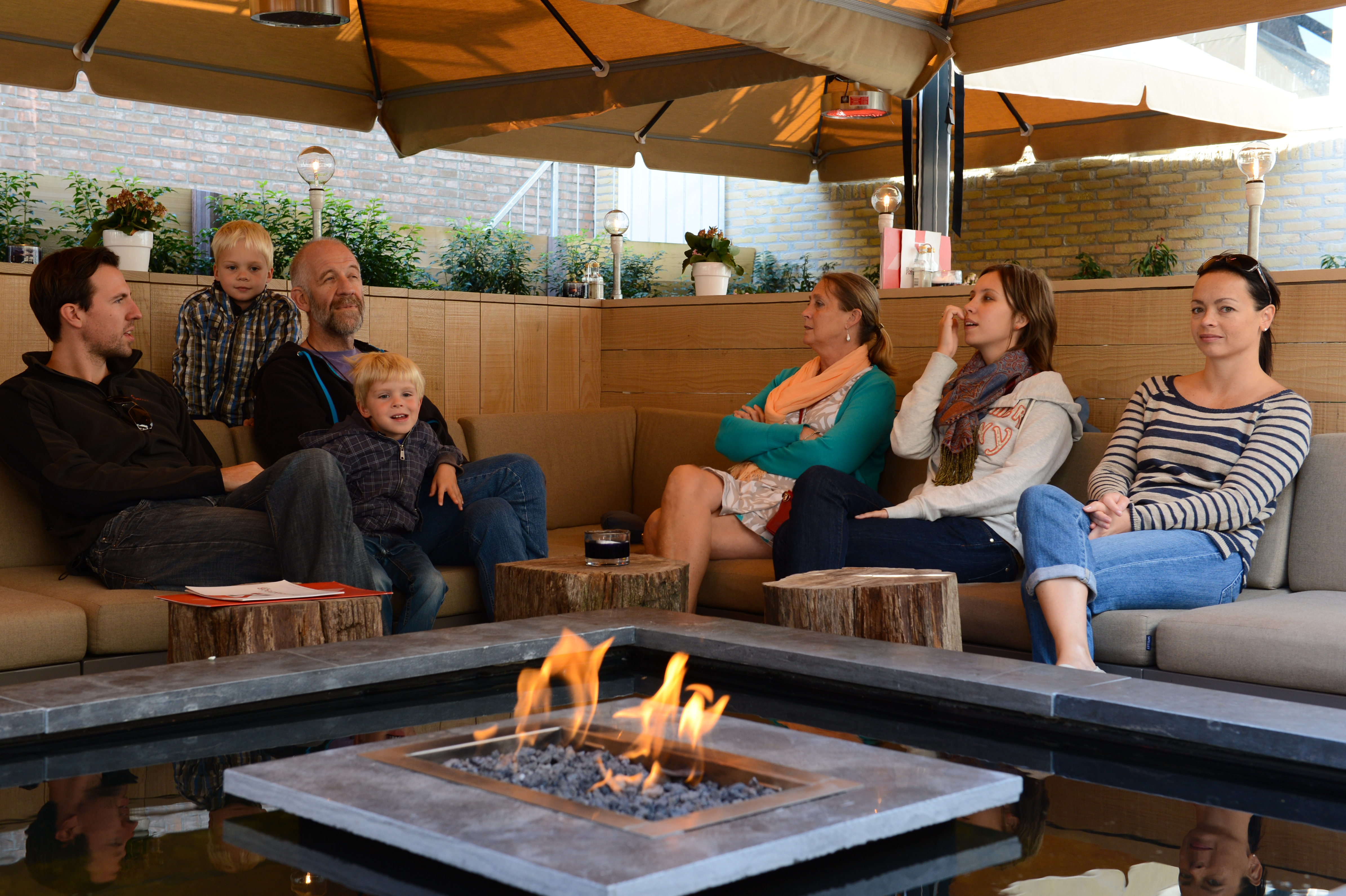 A group of people, including two children, sit around a fire pit under a covered outdoor seating area.