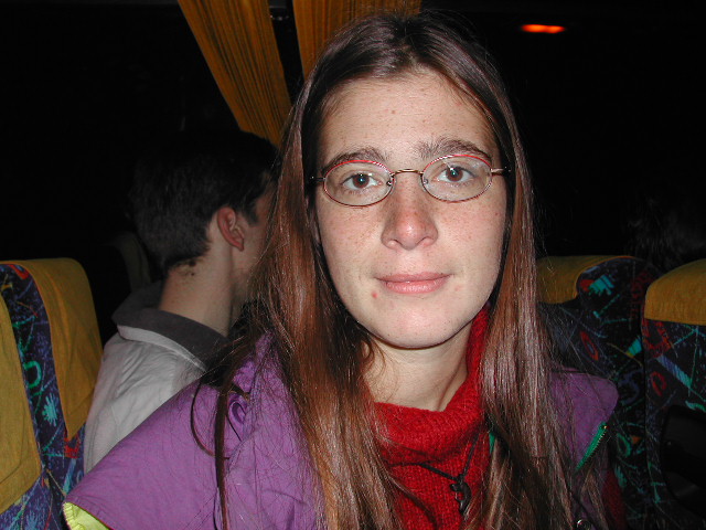 A woman with glasses and long brown hair sits on a bus, looking directly at the camera.