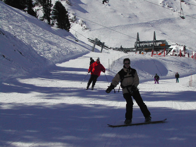 A person snowboarding down a ski slope while others ski behind them on a sunny winter day.