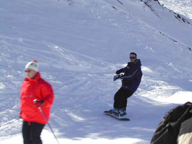 A person snowboards down a snowy slope while another person in a red jacket walks with ski poles.
