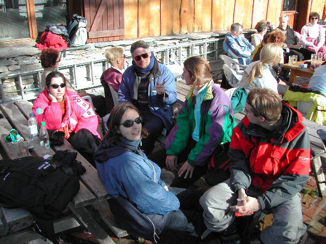 A group of people in winter clothing sit outdoors on wooden benches, talking and drinking in the sunlight.