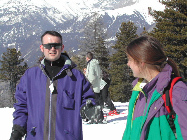 A man in a purple jacket talks while a woman in a colorful jacket listens on a snowy mountain.