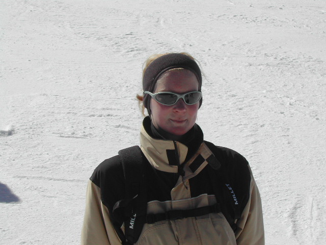 A person wearing sunglasses and a winter jacket stands on a snowy surface, looking at the camera.