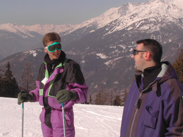 Two people in ski gear stand on a snowy slope, smiling and talking with mountains in the background.