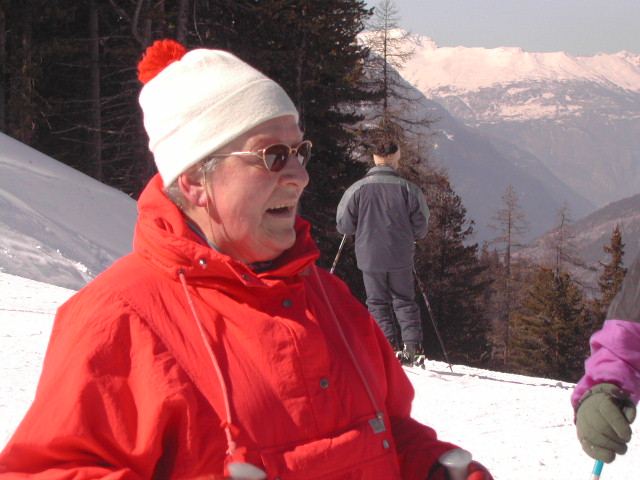 A person in a red jacket and white hat smiles while skiing on a snowy mountain with others in the background.
