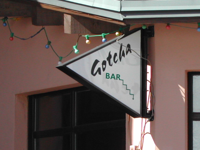 A triangular sign reading "Gotcha Bar" hangs on a building with string lights above it.