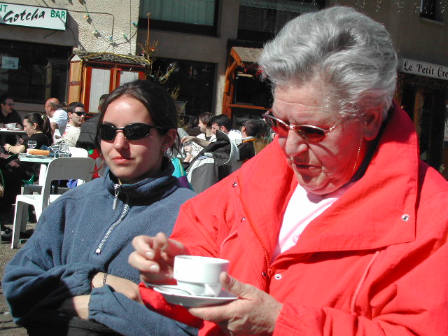 An older woman in a red jacket stirs her coffee, while a younger woman in sunglasses sits beside her.