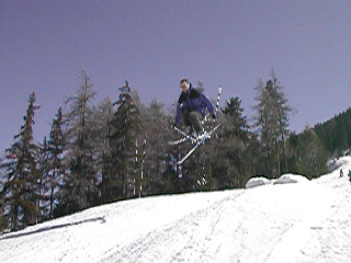 A skier in mid-air after jumping off a snowy slope, with trees in the background.