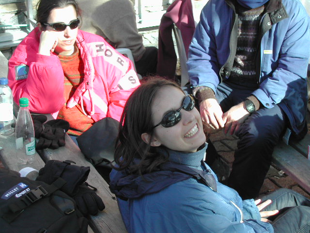 A smiling person in a blue jacket and sunglasses sits at an outdoor table with two others wearing winter clothing.