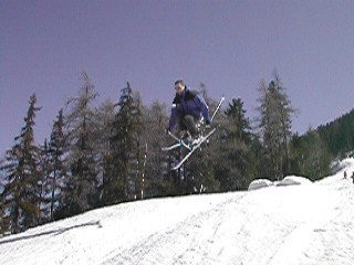 A skier in a blue jacket jumps off a snowy slope, holding skis in the air.
