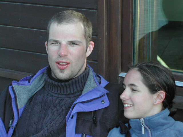 A man and a woman in winter jackets sit close together, with the woman smiling and looking at him.